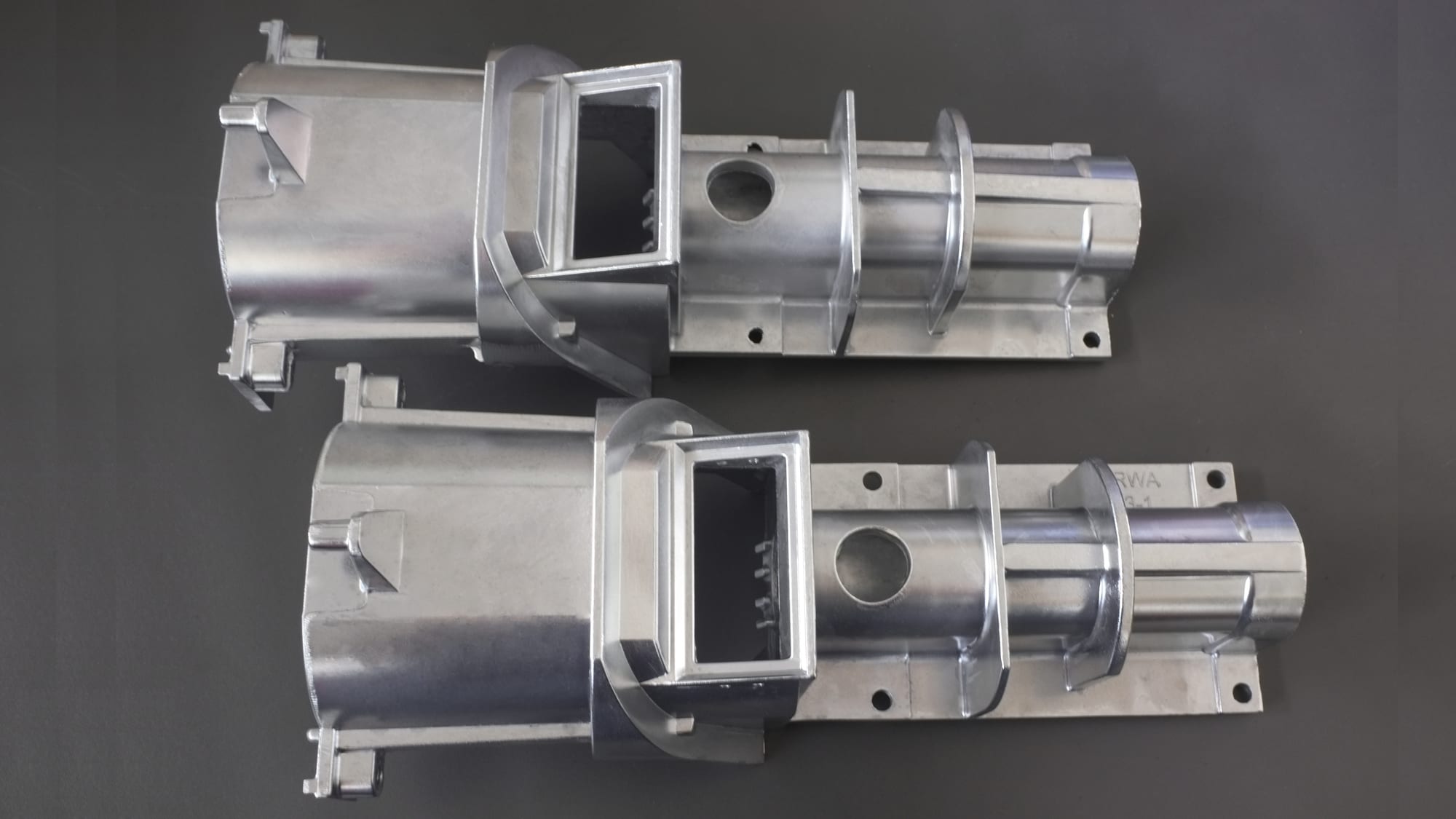 The image shows a pair of aluminum die casting parts on a gray background. The parts are made of metal and have a metallic sheen. They are arranged in a symmetrical pattern, with the largest part in t